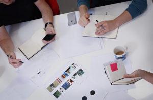 Ihmisiä suunnittelupöydän ääressä käsissään kynät ja vihot. Pöydällä suunnittelupapereita.